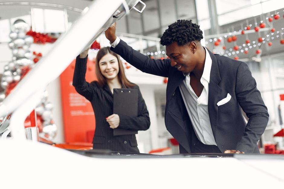 A customer and car salesperson sitting across a desk reviewing paperwork in a dealership showroom