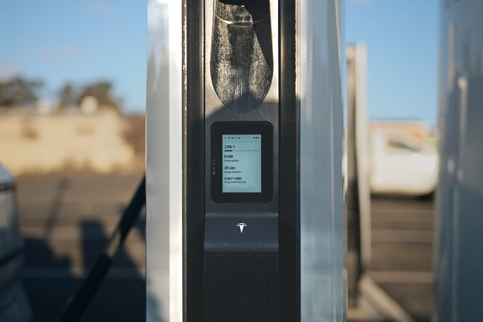 A hand holding a smartphone displaying a charging network app next to a public EV charging station